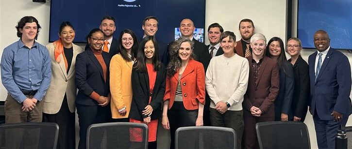 Group photo of a diverse team of professionals standing in a conference room. The group includes 16 individuals, a mix of men and women, dressed in business and business-casual attire. They are positioned in two rows, smiling for the camera. The background features a presentation screen with visible text, though not clearly readable in the image. The atmosphere appears professional and collaborative